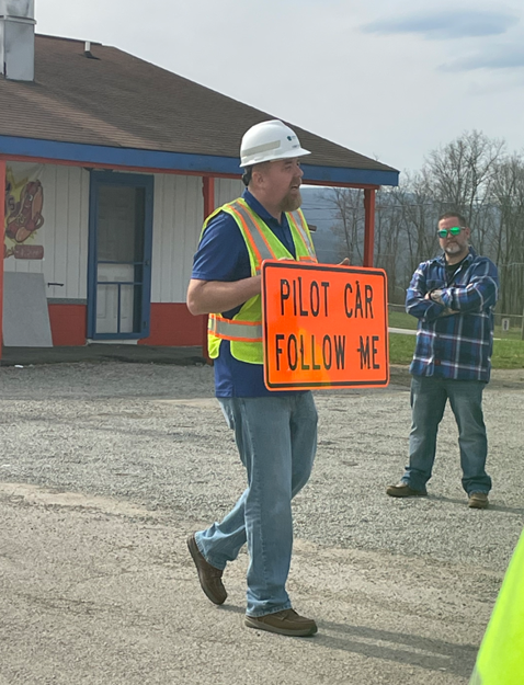 An image of an individual in a yellow safety vest holding an orange sign that says Pilot Car Follow Me